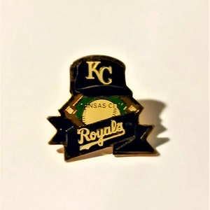 May include: A Kansas City Royals baseball team pin. The pin features a navy blue baseball cap with "KC" in gold lettering. Below the cap is a banner with the word "Royals" in gold lettering on a navy blue background.
