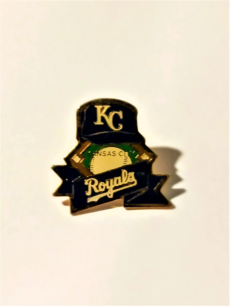 May include: A Kansas City Royals baseball team pin. The pin features a navy blue baseball cap with "KC" in gold lettering. Below the cap is a banner with the word "Royals" in gold lettering on a navy blue background.