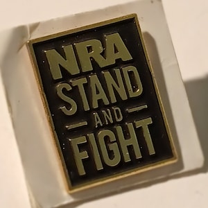 Puede incluir: Un pin de metal dorado con fondo negro. El pin dice "NRA Stand and Fight" en letras doradas.