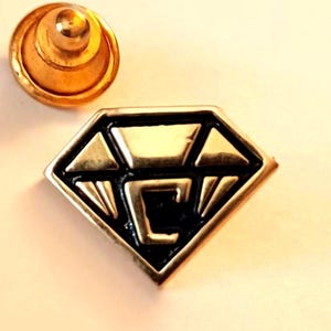 May include: A gold-toned diamond-shaped pin with a black geometric design in the center. The pin has a gold-toned back with a safety pin closure.