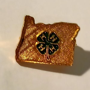 May include: A gold-colored pin in the shape of the state of Oregon. The pin features a textured surface and a green and black four-leaf clover emblem in the center. The pin is likely a souvenir or collectible item.