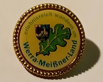 May include: A gold-colored pin with a yellow background and a green oak leaf. The text around the edge reads "...erlebnisreich wandern im Wera-Meißner-Land".