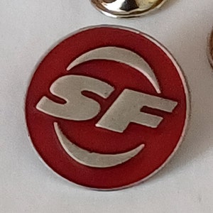 Puede incluir: Un pin circular rojo con un logotipo 'SF' plateado dentro de una media luna.