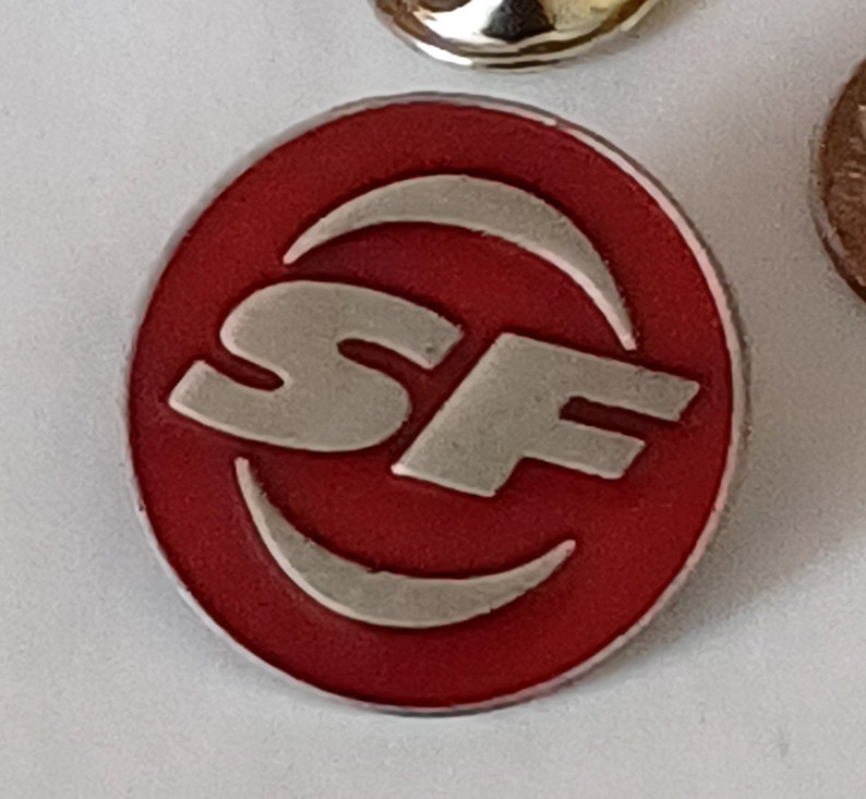 K&ouml;nnte beinhalten: Ein roter runder Pin mit einem silbernen 'SF'-Logo in einem Halbmond.