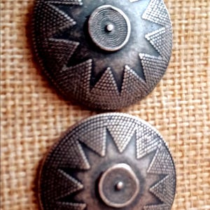 May include: Two antique silver-toned metal buttons. Each button is round with a central raised circle and a surrounding starburst design. The buttons have a textured surface and are set against a neutral woven background.