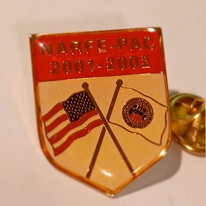 Op de afbeelding: Een gouden speld met een rode schildvormige achtergrond. Het schild heeft de tekst "NARFE-PAC 2001-2002" in gouden letters. Het schild heeft ook twee gekruiste vlaggen, een Amerikaanse en een met een gouden zegel.