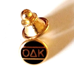 May include: Gold-colored pin with a black circle and the letters "OAK" in white. The pin has a gold-colored back with a ball on top.