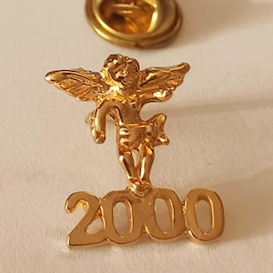 Op de afbeelding: Een goudkleurige speld met een cherubijn met uitgespreide vleugels, boven het getal "2000". De speld is waarschijnlijk gemaakt van metaal en heeft een glanzende afwerking. Een andere goudkleurige speld is zichtbaar op de achtergrond.