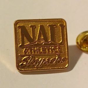 Puede incluir: Pin de color dorado con el texto "NAU Athletics Lumberjacks" en forma cuadrada.