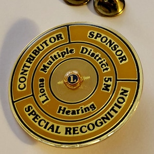 May include: A gold-colored Lions Club pin with the words "CONTRIBUTOR," "SPONSOR," and "SPECIAL RECOGNITION" in black text. The pin also features the words "Lions Multiple District 5M Hearing" and a small emblem with the letter "L."