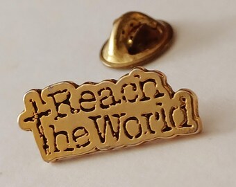 May include: A gold-colored lapel pin with the words "Reach the World" in a stylized font. The pin has a rectangular shape with a slightly irregular border. A separate gold-colored pin back is also visible.