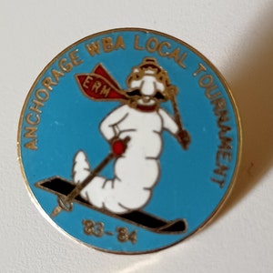 Puede incluir: Un pin de oro con fondo azul y un gusano blanco de dibujos animados esquiando. El gusano lleva un gorro rojo y blanco y una bufanda roja con las letras "ERM" en ella. El pin dice "Anchorage WBA Local Tournament 83-84".
