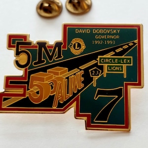 May include: A gold-colored pin with the text "5M 55 ALIVE 7" and the words "David Dobovsky Governor 1992-1993" on a green background. The pin also features a road design and the words "Circle-Lex Lions". Two gold-colored pins are in the background.
