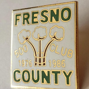 May include: A rectangular enamel pin with a white background and gold trim. It features the green text FRESNO COUNTY, the gold text 500 CLUB, the years 1976 and 1986, and a central graphic of three cotton bolls on stems in gold and brown.