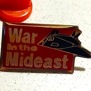 May include: A red and gold pin with the text "War in the Mideast" and a black silhouette of a stealth bomber.