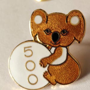 May include: A gold-toned metal enamel pin shaped like a koala bear. The koala has textured brown fur, a black nose, and white inner ears. It is holding a large white circular disc featuring the number 500 in gold outline font on its left side.