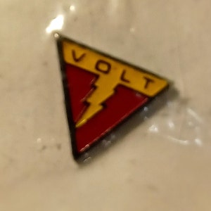 May include: A red and yellow triangular pin with a lightning bolt and the word "VOLT" in yellow letters.
