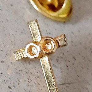 May include: A gold-colored cross pin with three round accents. The pin has a textured surface and is attached to a gold-colored backing.