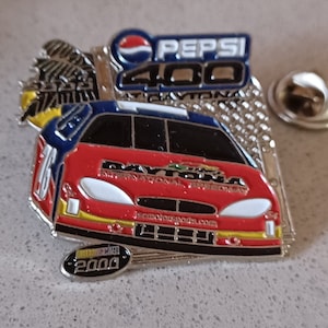 May include: A red, white, and blue NASCAR race car pin with the Pepsi 400 logo and the Daytona International Speedway logo. The pin is silver and has a 2000 date on it.