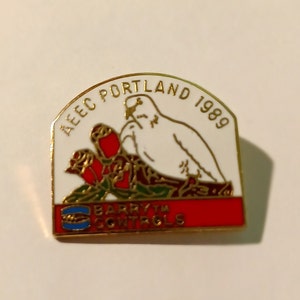 May include: A gold-colored pin with a white background. The pin features a white dove with red roses and green leaves. The text "AEEC PORTLAND 1989" is written in a gold arc above the dove. The text "BARRYTH CONTROLS" is written in gold on a red and blue stripe at the bottom of the pin.