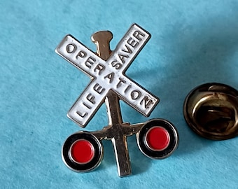 May include: A silver and gold Operation Lifesaver railroad crossing sign pin with red and black circles. The sign reads "Operation Lifesaver" and "Life"