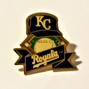 May include: A gold-toned pin featuring the Kansas City Royals baseball team logo. The pin includes a navy blue baseball cap with "KC" in gold, a baseball, and a banner with "Royals" in white script. The pin has a gold border.