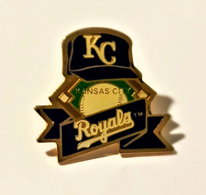 May include: A gold-toned pin featuring the Kansas City Royals baseball team logo. The pin includes a navy blue baseball cap with "KC" in gold, a baseball, and a banner with "Royals" in white script. The pin has a gold border.