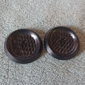 May include: Two dark brown, round buttons with a diamond pattern on the face and two holes for sewing. The buttons have a slightly raised edge and are set against a textured, light gray background.