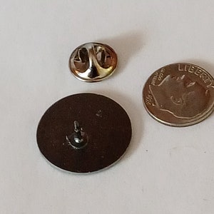 K&ouml;nnte beinhalten: Die R&uuml;ckseite einer runden Anstecknadel mit silberner Oberfl&auml;che. Die Nadel hat einen Sicherheitsverschluss auf der R&uuml;ckseite. Die Nadel ist etwa so gro&szlig; wie eine deutsche Zehn-Cent-M&uuml;nze.