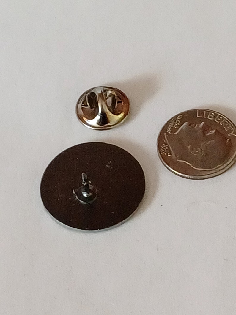 K&ouml;nnte beinhalten: Die R&uuml;ckseite einer runden Anstecknadel mit silberner Oberfl&auml;che. Die Nadel hat einen Sicherheitsverschluss auf der R&uuml;ckseite. Die Nadel ist etwa so gro&szlig; wie eine deutsche Zehn-Cent-M&uuml;nze.