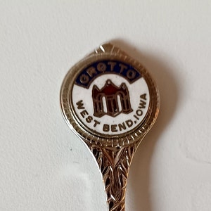 May include: A silver souvenir spoon with a blue and white enamel design. The design features a building with the text "GROTTO WEST BEND, IOWA".