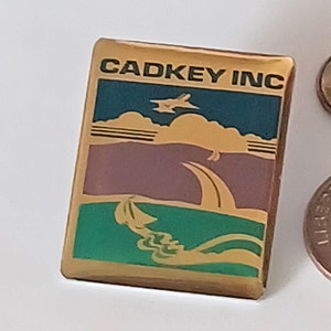 Pode incluir: Um alfinete dourado com uma imagem colorida de um avião voando sobre uma paisagem. O texto "CADKEY INC" está impresso em preto no topo do alfinete.