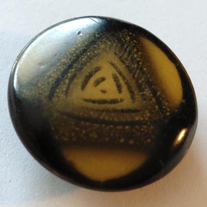 May include: A round, smooth button featuring a dark brown and amber-colored surface with a central triangular motif. The design consists of a smaller triangle nested within a larger one, rendered in a dark, etched-style line pattern.