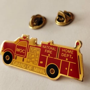 May include: A gold-toned metal lapel pin shaped like a fire truck with red enamel detailing. The pin features the text MOC, NATIONAL FIRE, HOME DEPT., and GRAND OF MINN. Two gold-toned metal clutch back fasteners are positioned above the pin.