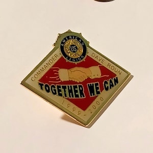 Op de afbeelding: Een gouden, ruitvormige pin met de woorden "Commander", "Dave Roun", "Together We Can" en "1999-2000". De pin heeft een handdruk en een rond embleem met de woorden "American Legion" en "US".