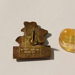 May include: A gold-colored pin with a detailed design of a flower. The back of the pin has the text "1987 J&E Inc. Atlanta, GA" printed on it.