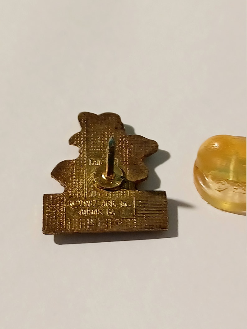 May include: A gold-colored pin with a detailed design of a flower. The back of the pin has the text "1987 J&E Inc. Atlanta, GA" printed on it.