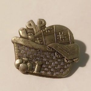Peut inclure: Une épingle en métal argenté en forme de panier rempli d'accessoires de couture. Le panier est tissé et contient une bobine de fil, des boutons et une paire de ciseaux.
