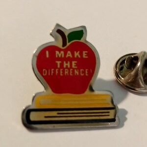 May include: A red apple with a green leaf and the text "I MAKE THE DIFFERENCE!" on it, sitting on top of a stack of yellow books. The apple and books are on a silver pin.