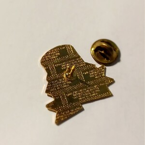 May include: A gold-colored pin with a detailed design, featuring a pin back and a separate, round clasp. The pin has a textured surface and a unique shape. The pin is on a white surface.