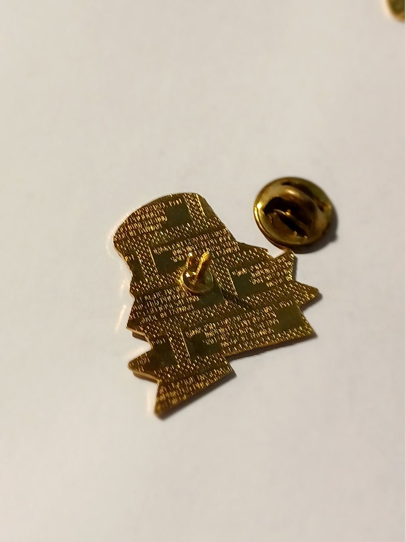 May include: A gold-colored pin with a detailed design, featuring a pin back and a separate, round clasp. The pin has a textured surface and a unique shape. The pin is on a white surface.