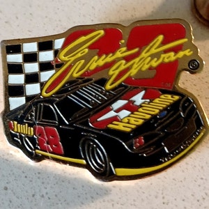 May include: A gold-colored pin featuring a black race car with a red number 28, a yellow and red "Havoline" logo, and a checkered flag. The pin also includes the signature "Ernie Irvan" in yellow lettering.
