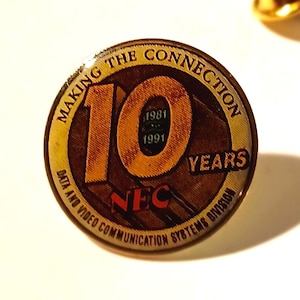 May include: A gold-coloured pin with a brown and yellow circular design. The centre features the number "10" with the years "1981" and "1991" inside. The text "Making the Connection" is above the number and "Years" is to the right. The text "NEC Data and Video Communication Systems Division" is below the number.