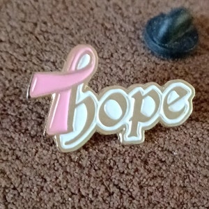 May include: Gold-tone pin with a pink ribbon and the word "Hope" in white lettering. The pin is on a brown fabric background.