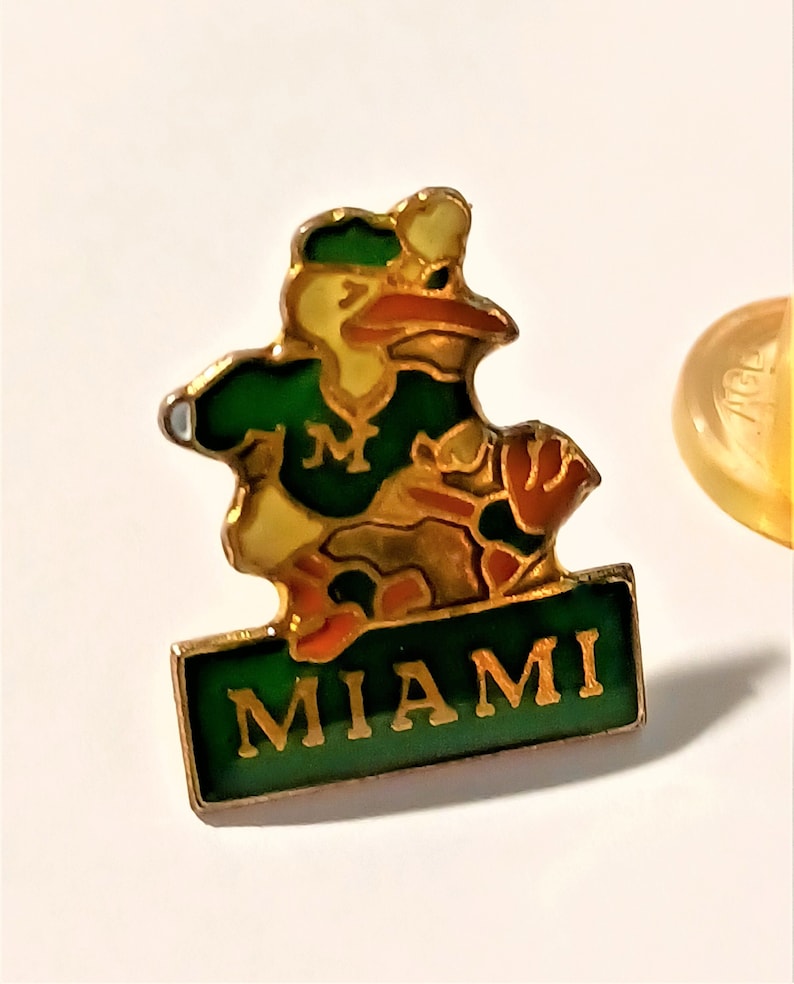 May include: A gold-colored pin with a cartoon bird wearing a green and yellow baseball uniform. The bird is holding a baseball glove and the pin says "MIAMI" in gold letters on a green background.
