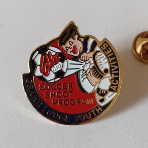May include: Gold pin with a cartoon character kicking a soccer ball. The pin says "Grand Lodge Youth Activities Soccer Shoot Program" and "USA" on the soccer ball.