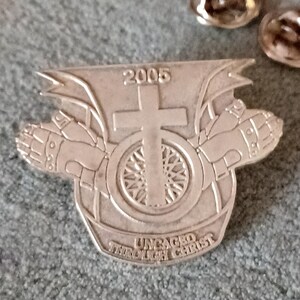 May include: Silver metal pin with a motorcycle wheel, cross, and two hands in prayer. The pin says "Uncaged Through Christ" and "2005".
