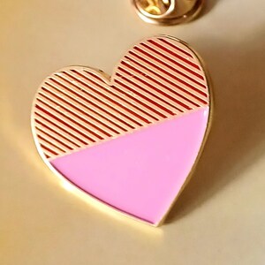 May include: A gold-tone heart-shaped pin with a pink and red striped design. The top half of the heart is red and white striped, and the bottom half is solid pink.