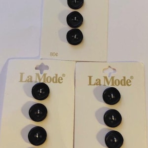 May include: Three cards of black La Mode brand buttons. Each card has a different number of buttons. The cards are cream-colored with the brand name printed in gold. The buttons are round with four holes.