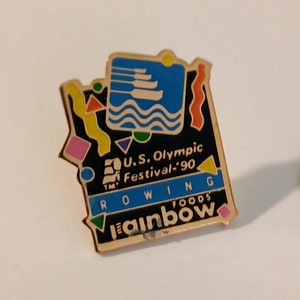 May include: A gold-coloured pin featuring a blue and white image of rowers in a boat. The pin has a black background with colourful geometric shapes and the text "U.S. Olympic Festival '90" and "ROWING" in white. The bottom of the pin says "Rainbow Foods" in gold.